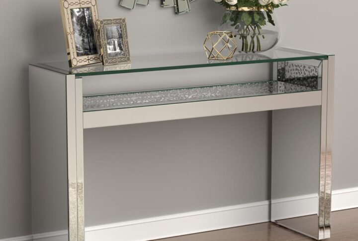 Keep a crisp linear décor theme with this console table. The clean lines form two square leg bases bridged by a shallow shelf with floating glass top above. The silver finish adds to the glamorous look of this striking piece. This silver console table adds a dramatic sculptural element to a living room or entertaining space. Top with a sculptural art piece for a truly captivating focal point.