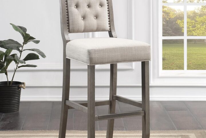 Elegant curves and a farmhouse style tufted back accented by bronze nail head trim make the Kamelin bar stool the perfect addition to your kitchen or dining space. Crafted of solid hardwood and finished in driftwood grey with a comfortable padded seat and back upholstered in durable and easy to clean beige polyester woven fabric.  Four-sided stretchers add strength and a comfortable foot rest with bronze metal cap to protect the wood finish.  Your purchase includes one (1) bar stool.